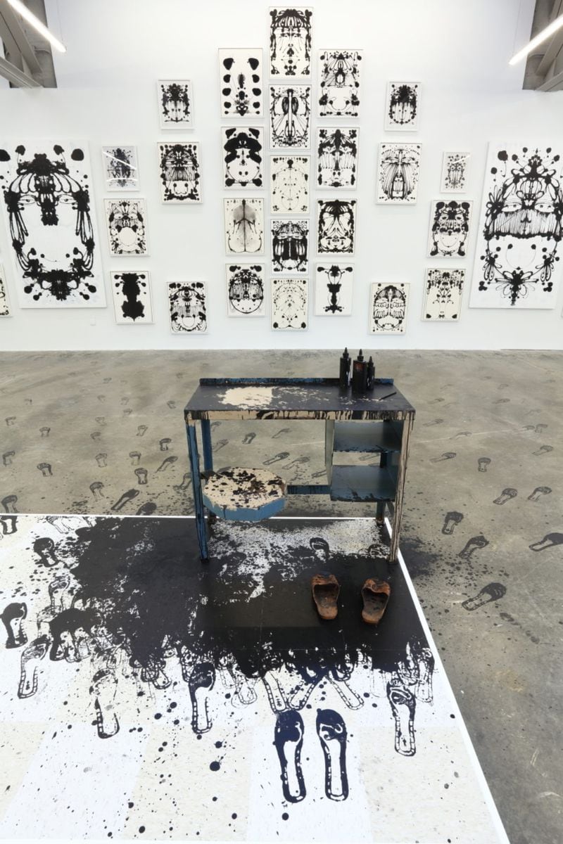 An installation view includes a desk with
Inky footprints leading to the centerpiece of the exhibit, a sprawling collection of unconventional black and white inkblots that form their own type of congruent blot in the way that they are arranged.