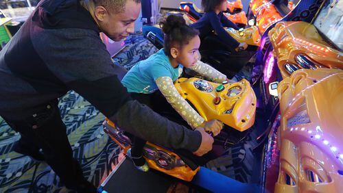 Muhammad Stewart (from left) helps his daughters Sophia Louann, 4, and Olivia Rose, 5, with their motorcycle adventure during a recent family outing at Andretti Indoor Karting and Games in Marietta. CURTIS COMPTON / CCOMPTON@AJC.COM