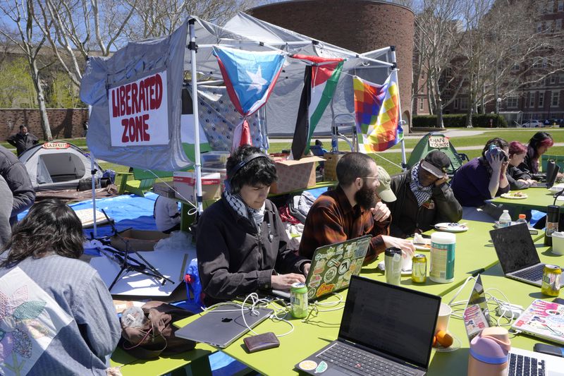 A Massachusetts Institute of Technology student, center, does school work on his computer while seated with others, Monday, April 22, 2024, at an encampment of tents on the MIT campus, in Cambridge, Mass. Students at MIT set up the encampment to protest what they said was MIT's failure to call for an immediate ceasefire in Gaza and to cut ties to Israel's military. (AP Photo/Steven Senne)