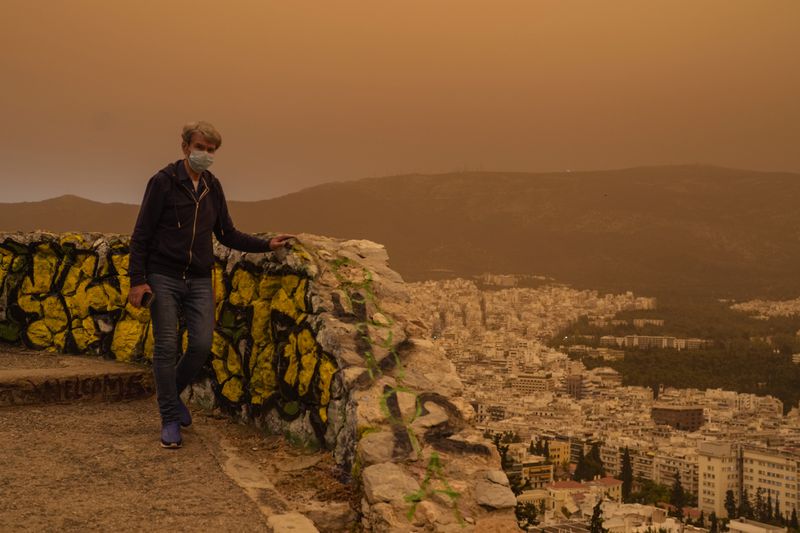 A man wearing a mask walks at the Lycabettus hill as the city of Athens is seen at the background, on Tuesday, April 23, 2024. The Acropolis and other Athens landmarks took on Martian hues Tuesday as stifling dust clouds blown across the Mediterranean Sea from North Africa engulfed the Greek capital. (AP Photo/Petros Giannakouris)
