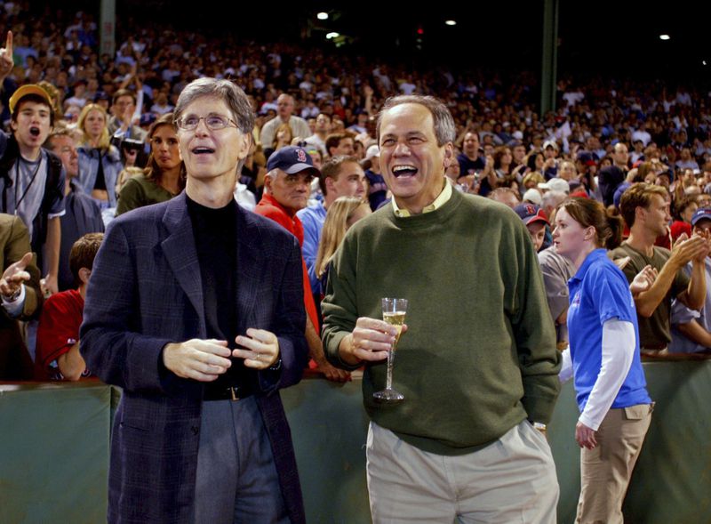 FILE - Boston Red Sox owners John Henry, left, and Larry Lucchino watch their team celebrate on the field after winning the American League wild-card at Fenway Park in Boston, Thursday, Sept. 25, 2003. Larry Lucchino, the force behind baseball’s retro ballpark revolution and the transformation of the Boston Red Sox from cursed losers to World Series champions, has died. He was 78. Lucchino had suffered from cancer. The Triple-A Worcester Red Sox, his last project in a career that also included three major league baseball franchises and one in the NFL, confirmed his death on Tuesday, April 2, 2024.(AP Photo/Winslow Townson)