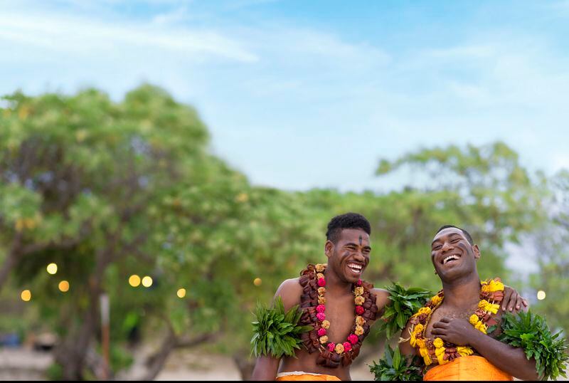 Two Meke men laughing on the beach. 
Photo by Tourism Fiji