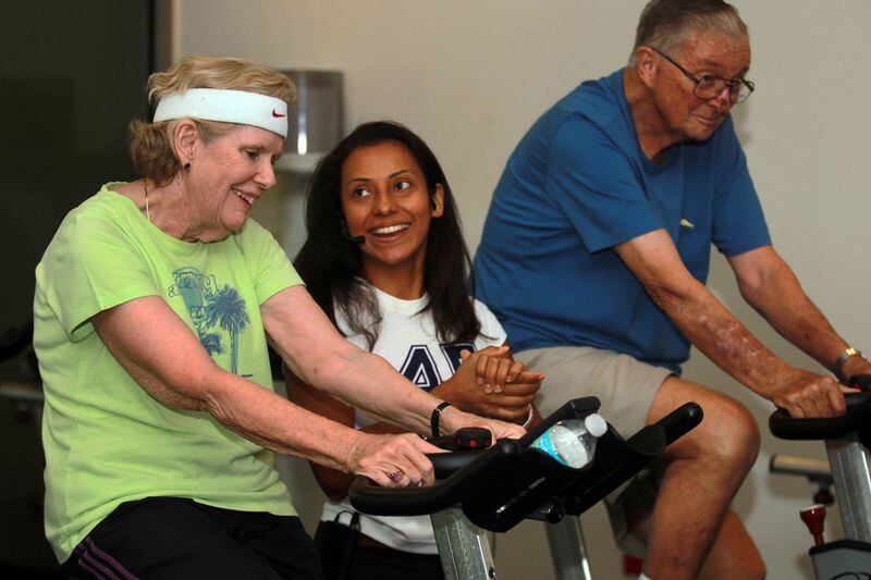 Angela Alvarado, a health coach instructor, helps Claire Hackett, left, and her husband Bob Hackett, right, during a cycling class for individuals with Parkinson's on Aug. 13, 2015.