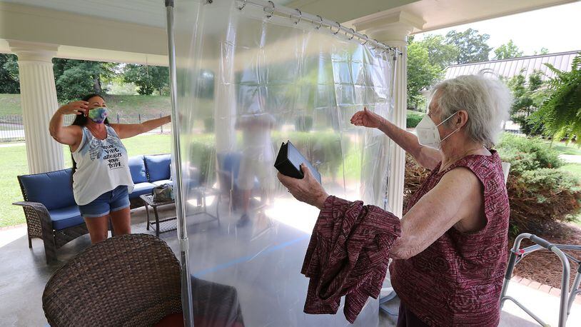 Elsie Grant, 86, sets her walker aside reaching for an air hug from her daughter Wanda Schroeder, socially distanced behind a protective plastic curtain when families could visit loved ones in the outdoor pavilion at Westbury Medical Care & Rehab earlier this month. CURTIS COMPTON / CCOMPTON@AJC.COM