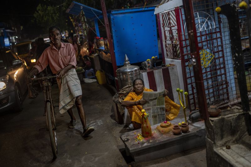 A man walks past as an elderly woman reading reads a newspaper outside a small temple by the street in the southern Indian city of Chennai, April 14, 2024. (AP Photo/Altaf Qadri)
