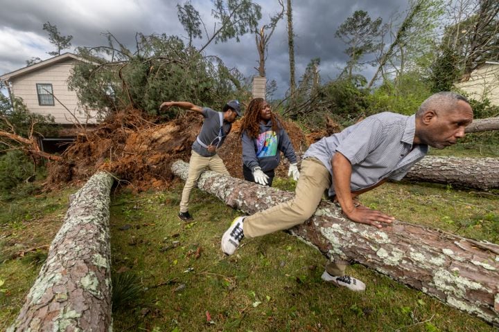 Laquan Grinter (from left), Dianne Carter and homeowner Terrance Harvell jump over downed trees at Harvell’s home on Briarwood Road on Wednesday, April 3, 2024. (John Spink / John.Spink@ajc.com)