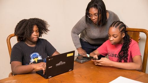 Online learning at the Hill house in Lithonia starts just after 5 pm when their mom gets home from her job at Lowe’s. Raina Hill, 12, left, a 6th grader at Champion Middle School logs in with help from her mother, LaTonya Hill, standing, while Rihanna Hill, 13, in red, an 8th grader at Champion Middle School uses a tablet to do school work. (Jenni Girtman for Atlanta Journal Constitution)