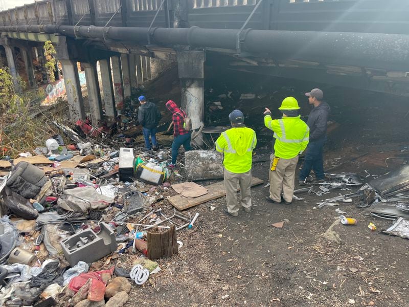 City employees and a cleanup crew inspect the remains of a homeless encampment built under a bridge on Cheshire Bridge. The encampment caught on fire last week, causing the closure of the road.