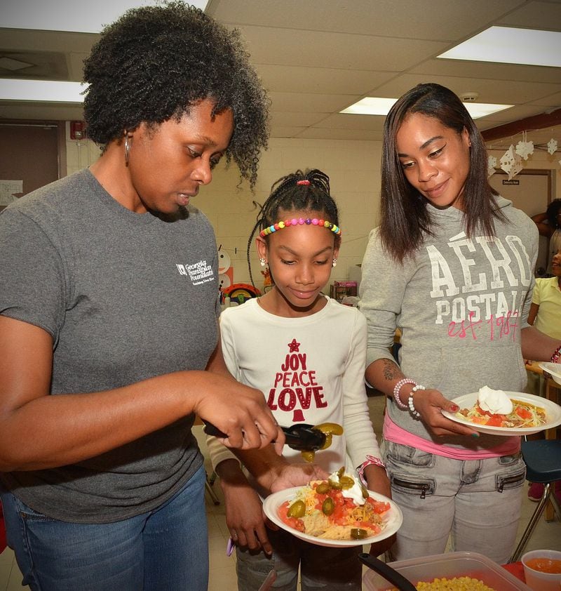Rosalind Watkins (left), a volunteer from Mount Zion AME Church, assists Laila Carter (center) with her plate full of tacos and peppers, while Laila’s mom, Ericka Carter, holds plates ready to serve the other children during dinnertime recently at Hagar’s House in Decatur. Hagar’s House, part of Decatur Cooperative Ministry, is an emergency shelter providing up to 90 days of shelter and support for families with children. Every night, volunteers arrive at Hagar’s House loaded with dinner. CHRIS HUNT / SPECIAL