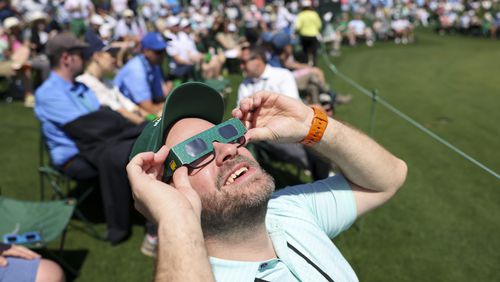 Tyler Steel, of Acworth, uses eclipse glasses to view the solar eclipse on the 18th green during the practice round of the 2024 Masters Tournament at Augusta National Golf Club, Monday, April 8, 2024, in Augusta, Ga. (Jason Getz / jason.getz@ajc.com)