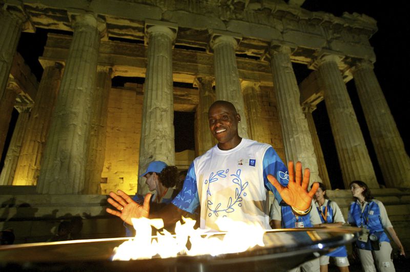 FILE - Nine times Olympic gold medal winner Carl Lewis of the United States poses for the photographers in front of the Olympic flame in front of the columns of the ancient Parthenon temple at the Acropolis hill, Aug. 12, 2004. On Tuesday, April 16, 2024 the flame for this summer's Paris Olympics will be lit and be carried through Greece for more than 5,000 kilometers (3,100 miles) before being handed over to French organizers at the Athens site of the first modern Olympics. (AP Photo/Lefteris Pitarakis, File)