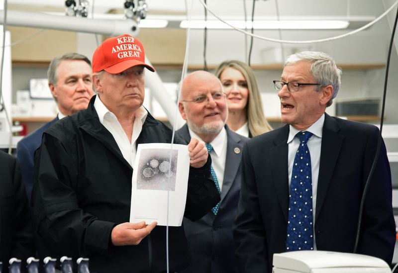 During his March tour of the CDC, President Trump held up a photograph of the coronavirus while the CDC’s Dr. Steve Monroe spoke to reporters. When the president made a false statement about the availabliity of tests at the time, CDC Director Robert Redfield, pictured at Trump’s right, did not correct him. HYOSUB SHIN / HYOSUB.SHIN@AJC.COM
