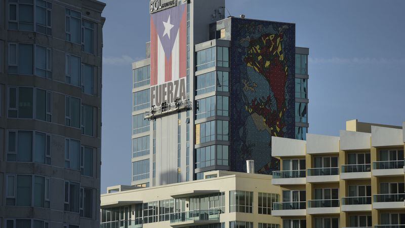 In this Friday, Oct. 20, 2017 photo, a crew hang a banner on the Caribe Hilton hotel featuring the Puerto Rican flag and the Spanish word "Strength," one month after Hurricane Maria in San Juan, Puerto Rico. The Caribe Hilton isn't accepting reservations until New Year's. (AP Photo/Carlos Giusti)