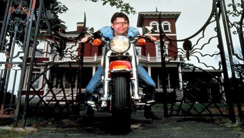Novelist Stephen King sitting on a motorcycle, at the gates of his house.