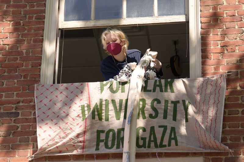 An activist uses keffiyeh scarves tied together to hoist a banner through an upper floor window of a Rhode Island School of Design building they have partially taken over Tuesday, May 7, 2024, at RISD, in Providence, R.I. The students and supporters are demanding that RISD condemn Israel's war effort in Gaza, and that the school divest from investments that benefit Israel. (AP Photo/Steven Senne)