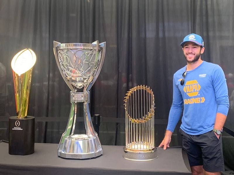 NASCAR driver Chase Elliott of Dawsonville took a tour of the College Football Hall of Fame on Wednesday in Atlanta. Here he stands with the trophies (from left) from the Georgia Bulldogs' 2021 College Football Playoff championship, the 2020 NASCAR Cup Series title won by Elliott and the 2021 World Series trophy, won by the Atlanta Braves. (Photo by Gabriel Burns/AJC)