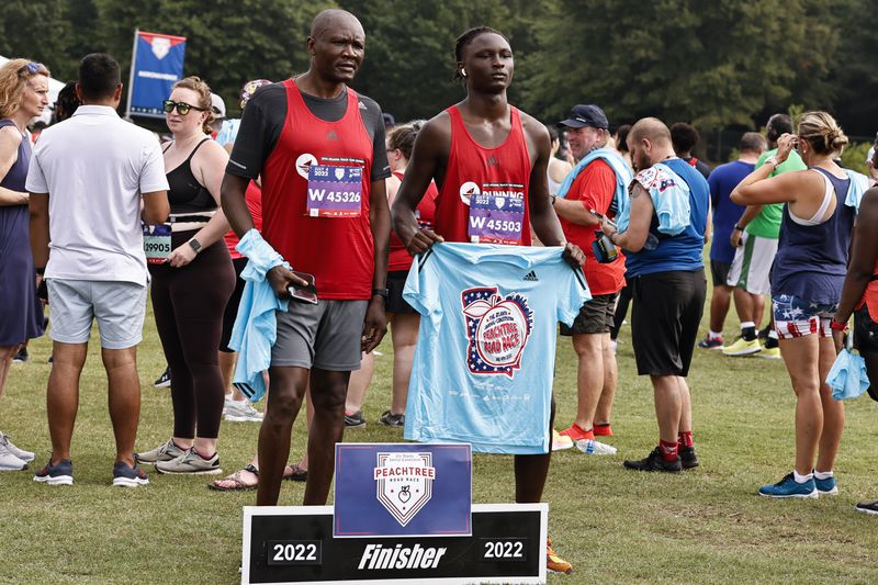 Dan Adhiambo (left) and his son James pose with  Peachtree Road Race t-shirts at Piedmont Park on Monday, July 4, 2022 (Natrice Miller/natrice.miller@ajc.com)