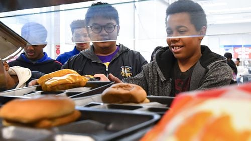 Eighth grader Elijah Caraballo, 14, reaches for a tray next to the hamburgers and Cheetos, as Alexander Boualavong (left), 15, waits his turn during lunch in the cafeteria at Elite Scholars Academy in Jonesboro. The school is in Clayton County, where all students get a free lunch so there is no meal debt. JOHN AMIS / FOR THE AJC