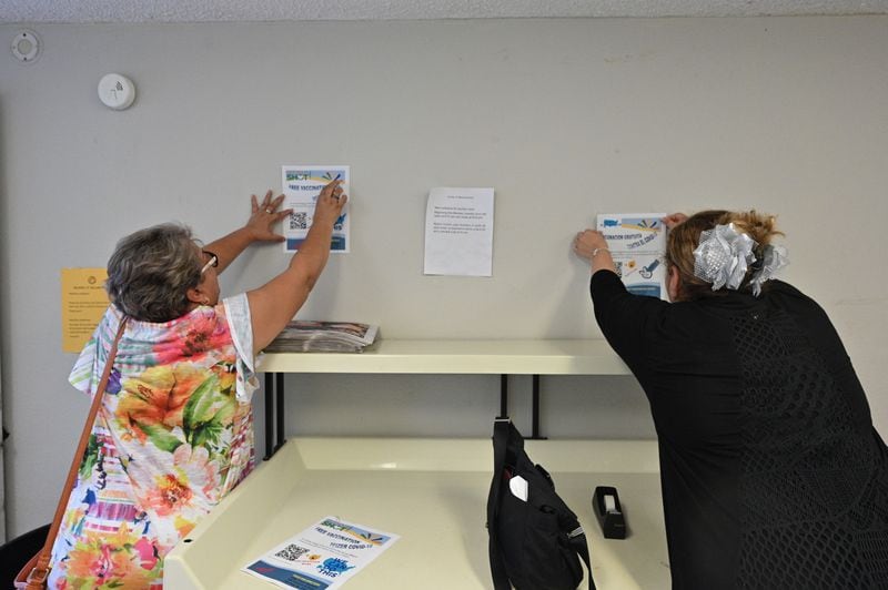 The Rev. Irma Guerra (right) and Aracely Hernandez put up COVID-19 vaccine pamphlets in a laundry room at Bloom at Meadowood Apartment Homes in Norcross on Wednesday, Aug. 11, 2021. Hernandez is a resident there and a member of Christ Church Episcopal. (Hyosub Shin / Hyosub.Shin@ajc.com)
