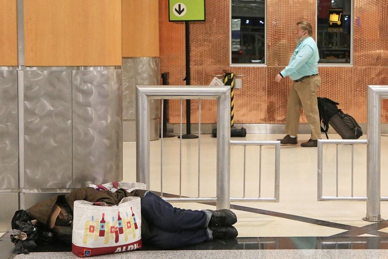 A man (left) sleeps on the ground in the domestic terminal of Hartsfield-Jackson International Airport as flight passengers (right) walk toward security on Friday, February 7, 2020, in Atlanta. Many of Atlanta’s homeless sleep overnight in the airport’s domestic terminal when the city experiences frigid winter temperatures. The man (left) wished not to be identified. (Christina Matacotta/crmatacotta@gmail.com)