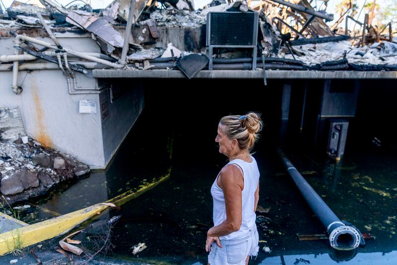 FILE - Janet Spreiter, whose home across the street was destroyed in the August wildfire, stands in front of a flooded parking garage in a destroyed business complex next to the Lahaina Shores Beach Resort on Front Street, Friday, Dec. 8, 2023, in Lahaina, Hawaii. The Maui Fire Department is expected to release a report Tuesday, April 16, 2024, detailing how the agency responded to a series of wildfires that burned on the island during a windstorm last August. (AP Photo/Lindsey Wasson, File)