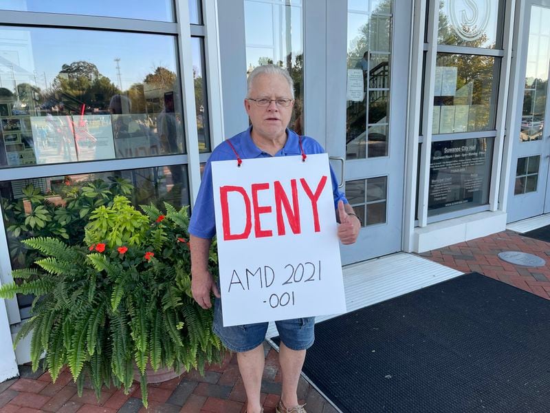 Mike Sutherland, a retired builder who has lived in Suwanee for 44 years, wears a "deny" sign draped over his shoulders outside Suwanee City Hall before City Council's Tuesday meeting. (Tyler Wilkins / tyler.wilkins@ajc.com)