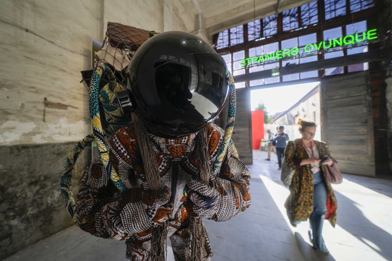 A visitor walks next to the installation 'Refugee Astronaut VIII' by artist Yinka Shonibare at the 60th Biennale of Arts exhibition in Venice, Italy, Tuesday, April 16, 2024. In background a neon write in Italian ''Straniero Ovunque'- Foreign Everywhere')'. The Venice Biennale contemporary art exhibition opens Saturday for its six-month run through Nov. 26. The main show titled Stranieri Ovunque – Foreigners Everywhere' is curated for the first time by a Latin American, Brazilian Adrian Pedrosa. Pedrosa is putting a focus on underrepresented artists from the global south, along with gay and Indigenous artists. Alongside the main exhibition, 88 national pavilions fan out from the traditional venue in Venice's Giardini, to the Arsenale and other locations scattered throughout the lagoon city. (AP Photo/Luca Bruno)