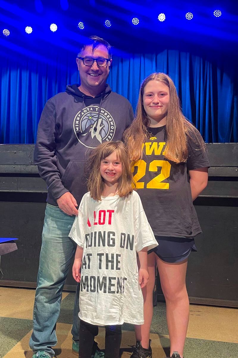 Mike Dean and his two daughters – one wearing a shirt that says "A LOT going on at the moment" in a nod to Taylor Swift, and the other wearing a shirt that says "Iowa 22" in reference to basketball star Caitlin Clark - smile inside the First Avenue concert venue Tuesday, May 7, 2024, after Gov. Tim Walz signed a so-called Taylor Swift bill. The law, prompted by the frustration a legislator felt at not being able to buy tickets to Swift's 2023 concert in Minneapolis, will require ticket sellers to disclose all fees up front and prohibit resellers from selling more than one copy of a ticket for live events held in Minnesota. (AP Photo/Trisha Ahmed)