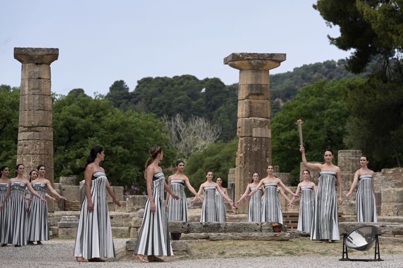 Actress Mary Mina, playing high priestess, right, holds a torch during the official ceremony of the flame lighting for the Paris Olympics, at the Ancient Olympia site, Greece, Tuesday, April 16, 2024. The flame will be carried through Greece for 11 days before being handed over to Paris organizers on April 26. (AP Photo/Thanassis Stavrakis)