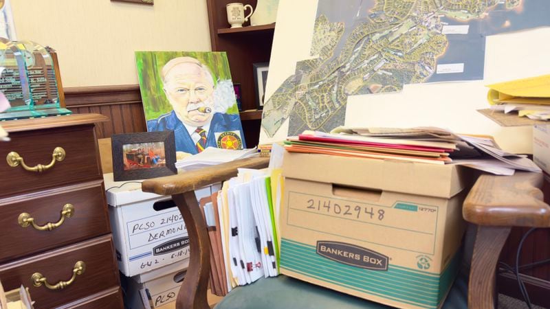 Stacks of Dermond-homicide files and other materials occupy a corner of Putnam County Sheriff Howard Sills' office in Eatonton. A painting of the sheriff is propped in the background. (Joe Kovac Jr. / AJC)