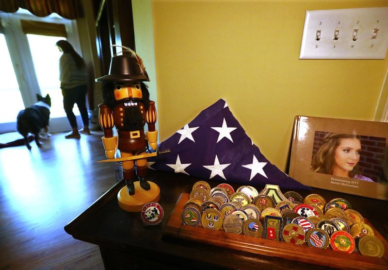 U.S. Army Col. David McCracken’s challenge coin collection and the American flag that covered his casket are on display beside a photo of his daughter, Mattie, in the family’s home in Tyrone. David’s wife, Tammy, and the family’s dog, Ranger, stand nearby. Curtis Compton/ccompton@ajc.com