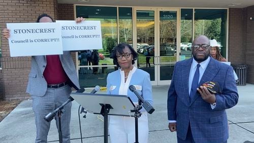 Michael Fayoyin, left, holds signs that read, "Stonecrest Council is Corrupt!!" during the news conference held by mayoral candidates Diane Adoma (center) and Kirby Frazier (right).