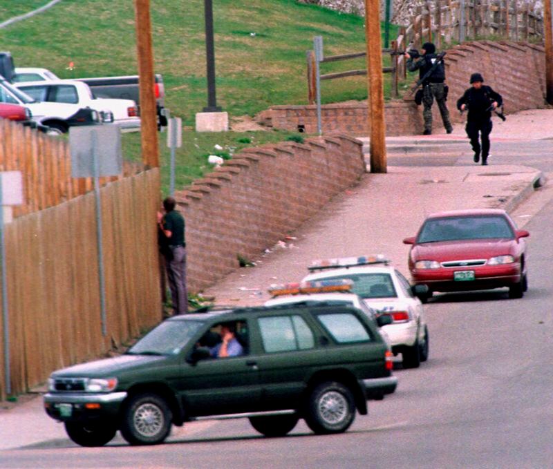FILE - SWAT members run down Pierce Street while a Jefferson County, Colo., Sheriff's Department deputy peers through a fence to keep an eye on Columbine High School after a pair of gunmen went on a shooting rampage inside the facility Tuesday, April 20, 1999, in the southwest Denver suburb of Littleton, Colo. (AP Photo/David Zalubowski, File)