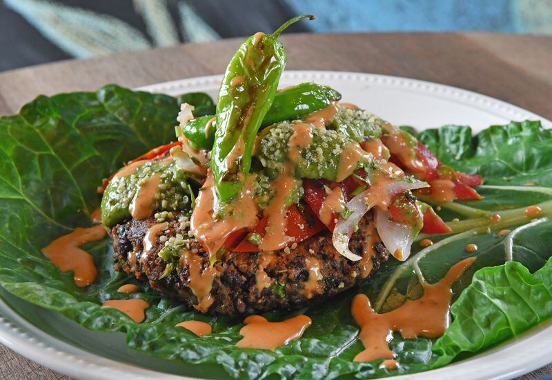 An Enchilada Lasagna Burger is served here on a blanched collard leaf, with Pico de Gallo, Chipotle Mayo, and Avocado Tomatillo Salsa garnished with blistered shishito peppers and hemp seeds. A hamburger bun is also a serving option. (Styling by Claudine Molson-Sellers / Chris Hunt for the AJC)