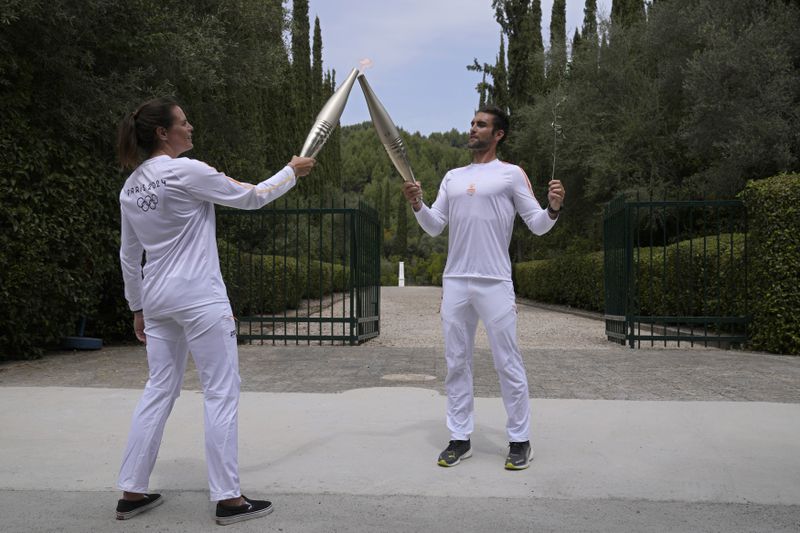 The first torch bearer, Greek olympic gold medalist Stefanos Douskos, right, passes the flame to first French torchbearer, three-time Olympic medallist Laure Manaudou, near the monument to Pierre de Coubertin, in the background, after the official ceremony of the flame lighting for the Paris Olympics, at the Ancient Olympia site, Greece, Tuesday, April 16, 2024. The flame will be carried through Greece for 11 days before being handed over to Paris organizers on April 26. (AP Photo/Thanassis Stavrakis)