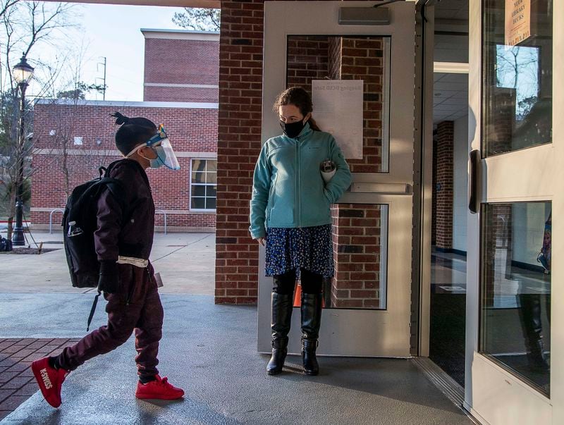 John R. Lewis Elementary School music teacher Elena Prestwood welcomes a student wearing a face shield and a face mask into the school building on the first day of in-person learning in March. (Alyssa Pointer / Alyssa.Pointer@ajc.com)