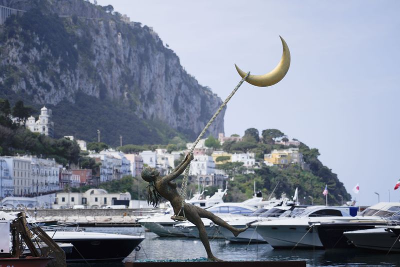 The bronze sculpture by Giacinto Bosco titled "swing on the moon" is backdropped by Capri's harbour, Wednesday, April 17, 2024. Group of Seven foreign ministers are meeting on the Italian resort island of Capri, with soaring tensions in the Mideast and Russia's continuing war in Ukraine topping the agenda. The meeting runs April 17-19. (AP Photo/Gregorio Borgia)