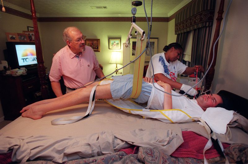 David Jayne is attended by his father, Bill, and his live-in caregiver Rose Delisser, during his normal morning routine which takes nearly two hours. A sling and hoist above his bed is used to lift him out of bed. (RICH ADDICKS / Staff)