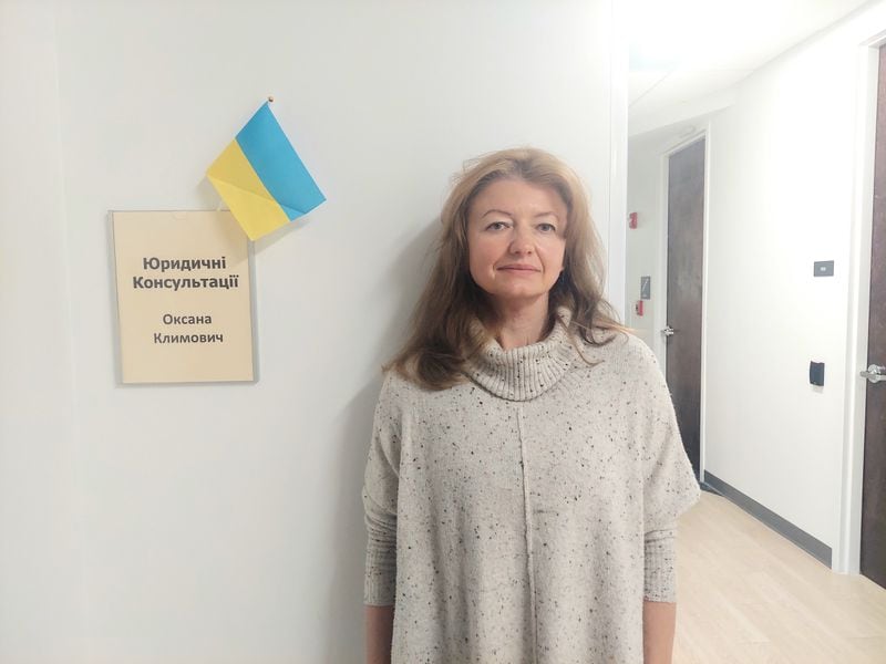 Oksana Klymovich, a volunteer with the Ukrainian Congress of America, stands next to the room where she provides free legal consultaitons for Ukrainian immigrants. She is standing next to a sign that says "legal consultations" in Ukrainian underneath a small Ukrainian flag. She has long brown hair parted down the middle, and is wearing a loose-fitting, beige, speckled turtleneck. The walls of the hallway she is standing in are white and illuminated by bright, flourescent, overhead lighting.