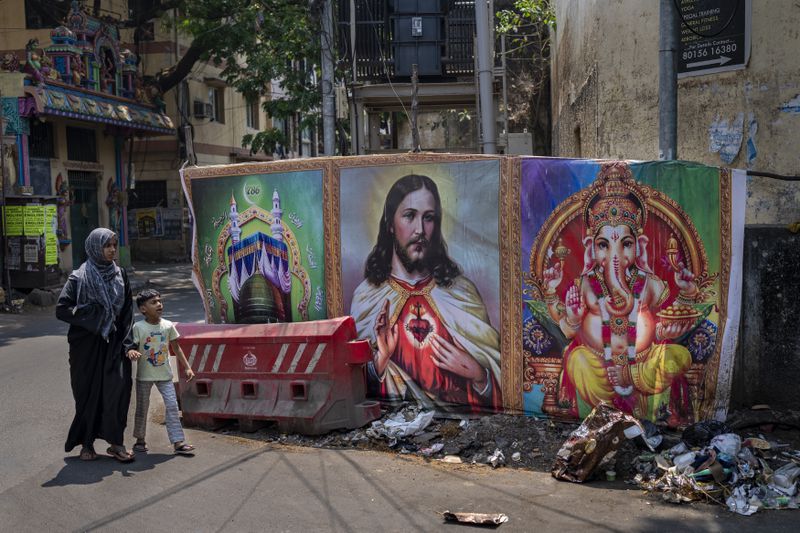 A Muslim woman and a boy walk past portraits of elephant-headed Hindu God Ganesha, Jesus Christ and Islam's holy sites of Mecca and Medina, in the southern Indian city of Chennai, April 18, 2024. (AP Photo/Altaf Qadri)