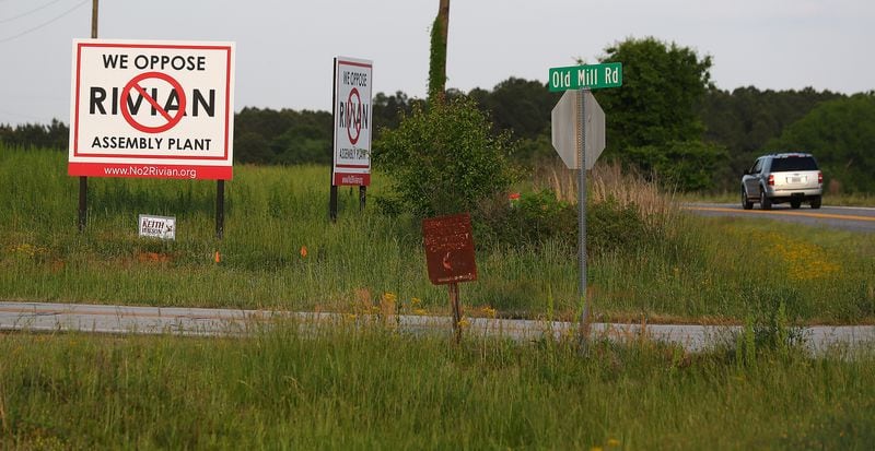 A motorist passes by large signs opposing the Rivian assembly plant at Old Mill Road and Davis Academy Road just across the intersection from the planned $5 billion Rivian electric vehicle plant on Monday, May 2, 2022, in Rutledge.  “Curtis Compton / Curtis.Compton@ajc.com”