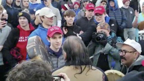 In this Friday, Jan. 18, 2019 image made from video provided by the Survival Media Agency, a teenager wearing a "Make America Great Again" hat, center left, stands in front of an elderly Native American singing and playing a drum in Washington. The Roman Catholic Diocese of Covington in Kentucky is looking into this and other videos that show youths, possibly from the diocese's all-male Covington Catholic High School, mocking Native Americans at a rally in Washington.