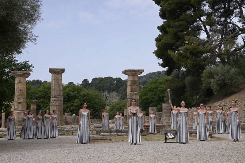 Actress Mary Mina, playing high priestess, right, holds a torch with the flame during the official ceremony of the flame lighting for the Paris Olympics, at the Ancient Olympia site, Greece, Tuesday, April 16, 2024. The flame will be carried through Greece for 11 days before being handed over to Paris organizers on April 26. (AP Photo/Thanassis Stavrakis)