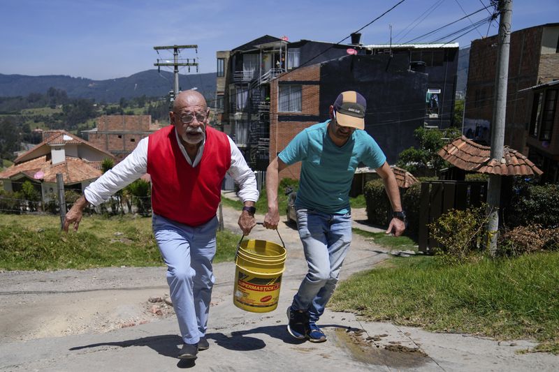 Residents carry a bucket of water to their home after filling it at a water distribution truck while water is rationed in La Calera, on the outskirts of Bogota, Colombia, Tuesday, April 16, 2024. Amid a drought linked to the El Niño weather pattern, several regions of Colombia have adopted measures to curb water consumption while reservoirs are low. (AP Photo/Fernando Vergara)