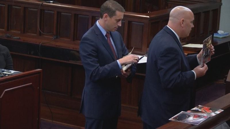 Detective Carey Grimstead (right) is assisted by prosecutor Jesse Evans as he shows the jury photos of Cooper's car seat during the murder trial of Justin Ross Harris at the Glynn County Courthouse in Brunswick, Ga., on Wednesday, Oct. 26, 2016. (screen capture via WSB-TV)