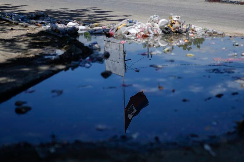 A Haitian flag is reflected in a puddle, lined with trash, in the Champ de Mars area of Port-au-Prince, Haiti, Tuesday, April 30, 2024. (AP Photo/Ramon Espinosa)