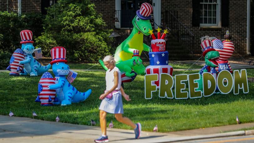 Alyson Cohen strolled by the display of dinosaurs on Dunwoody Club Drive near Braffington Court in Dunwoody on Friday, June 9, 2023. The display of dinosaurs first appeared in the neighborhood in 2020 then quickly evolved into holiday themed displays along with encouraging messages.  (John Spink / John.Spink@ajc.com)
