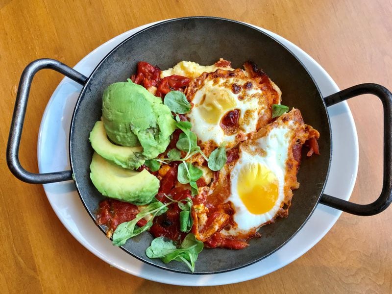 Southern Shakshuka at Tupelo Honey features two baked eggs in a Creole sauce over goat cheese grits. The dish is garnished with fresh avocado slices and lamb’s lettuce. LIGAYA FIGUERAS / LFIGUERAS@AJC.COM