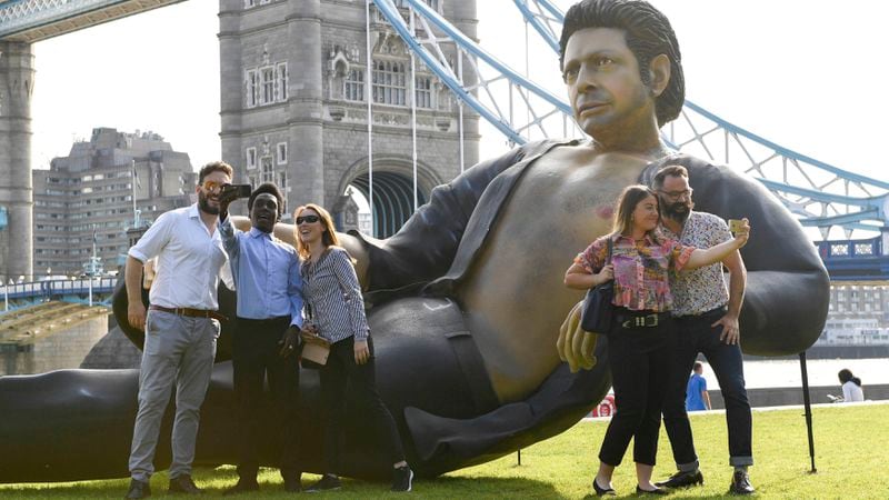 People take photos by a 25ft statue of actor Jeff Goldblum in a pose from a scene in the first Jurassic Park movie, which has been created by a TV channel to celebrate the film's 25th birthday, at Potters Fields Park, London, Wednesday July 18, 2018. Tower Bridge in the background.