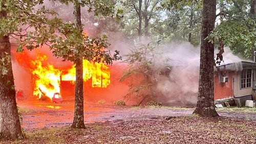 A one-story red and tan house is pictured on fire with flames blazing out of the front door and windows. Smoke is pouring out of the structure, and tall trees surround the home.